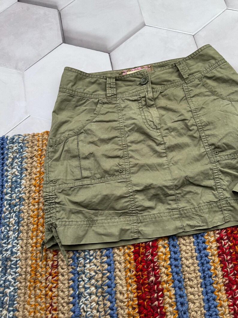 May include: A green cargo skirt with a button fastening and multiple pockets. The skirt has a drawstring at the bottom.
