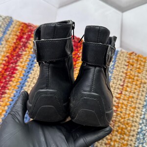 May include: Pair of black leather ankle boots with zippers and buckle straps. The boots have a rounded toe and a rubber sole. The boots are held by a gloved hand.