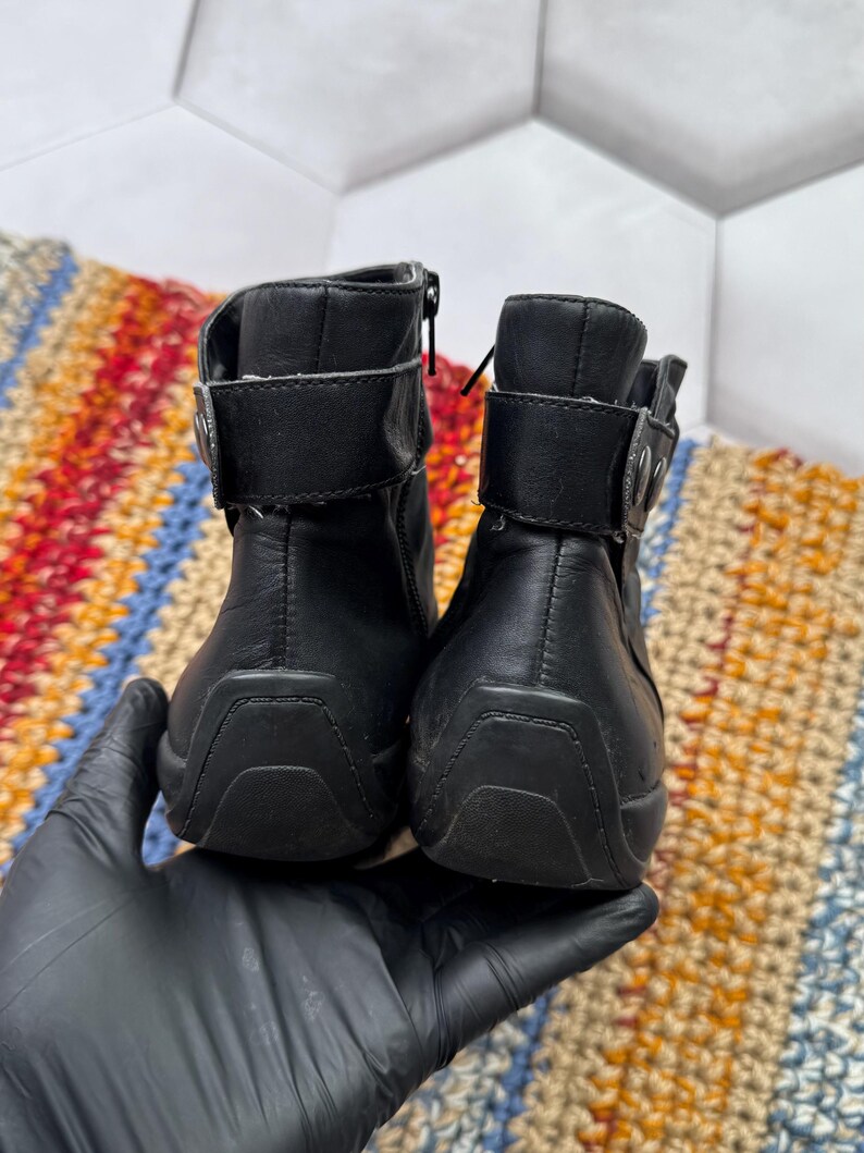 May include: Pair of black leather ankle boots with zippers and buckle straps. The boots have a rounded toe and a rubber sole. The boots are held by a gloved hand.
