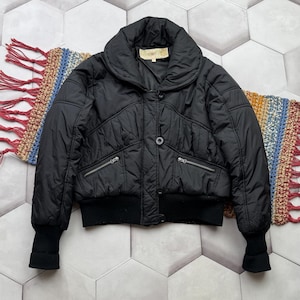 May include: Black quilted jacket with a wide collar, two zippered pockets, and a button closure. The jacket has a ribbed hem and cuffs. A colorful fringed textile is partially visible beneath the jacket.