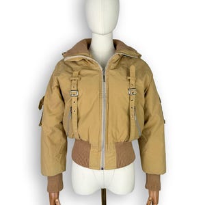 May include: A tan puffer jacket featuring a high collar, a full-length front zipper, and two vertical straps with silver-toned metal buckles on the chest. The jacket includes ribbed knit cuffs and a ribbed hem, with long sleeves and a cropped fit.