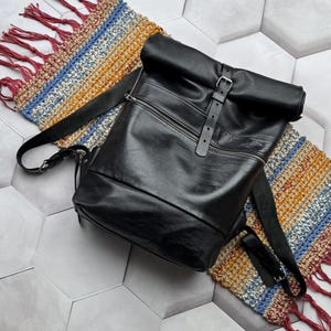 Peut inclure: Un sac à dos en cuir noir avec une fermeture à rabat et des bretelles réglables. Le sac à dos est posé sur un tapis rayé multicolore avec des franges.
