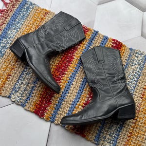 Könnte beinhalten: Ein Paar schwarze Cowboy-Stiefel mit weißen Stickereidetails, die auf einem farbenfrohen, gestreiften Teppich liegen. Die Stiefel haben ein klassisches Western-Design mit spitzer Zehenpartie und niedrigem Absatz. Der Teppich zeigt rote, blaue, gelbe und beige Streifen.