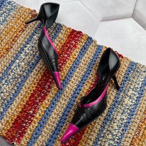 May include: A pair of black leather high heels with pink trim. The heels are pointed toe and have a thin strap across the top of the foot. The heels are sitting on a striped rug.