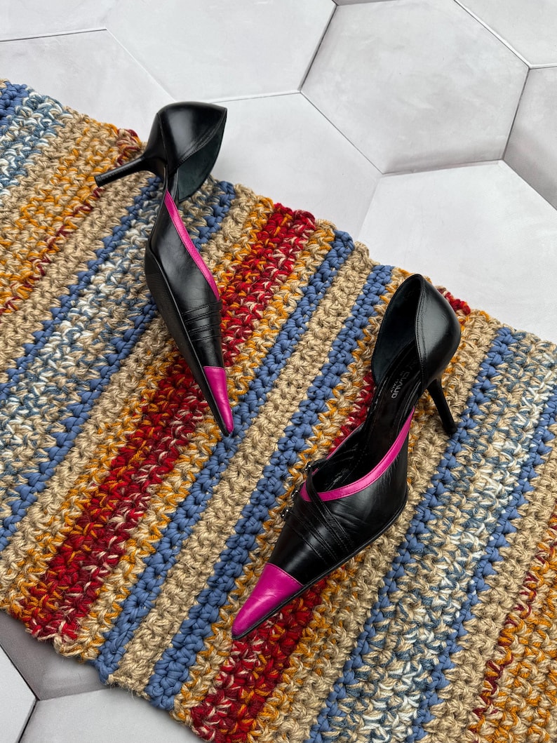 May include: A pair of black leather high heels with pink trim. The heels are pointed toe and have a thin strap across the top of the foot. The heels are sitting on a striped rug.