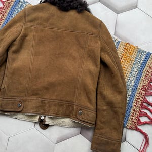 May include: Brown suede jacket with a black shearling collar. The jacket features a zip closure, two buttoned pockets, and a cream-colored lining. The back of the jacket is visible, showcasing the tailored seams and classic design.
