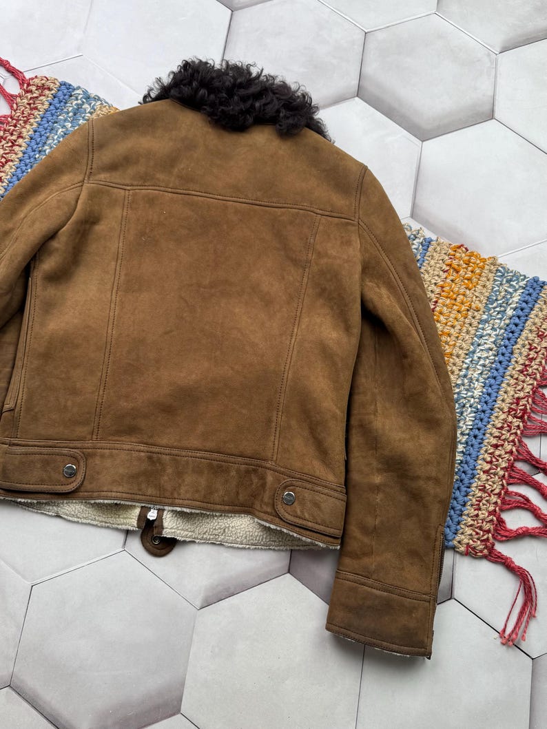 May include: Brown suede jacket with a black shearling collar. The jacket features a zip closure, two buttoned pockets, and a cream-colored lining. The back of the jacket is visible, showcasing the tailored seams and classic design.