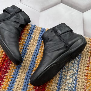 May include: Black leather ankle boots with side zippers and buckle straps. The boots are on a colorful, textured rug with blue, tan, and orange stripes. The boots have a low heel and a rounded toe.
