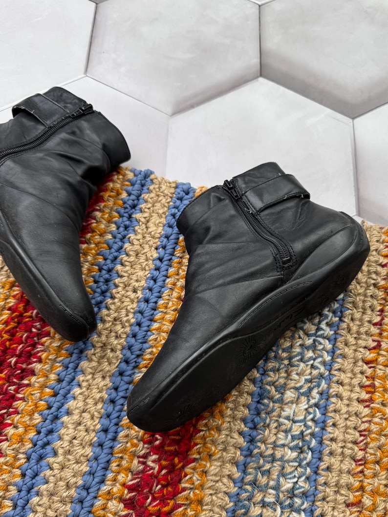 May include: Black leather ankle boots with side zippers and buckle straps. The boots are on a colorful, textured rug with blue, tan, and orange stripes. The boots have a low heel and a rounded toe.