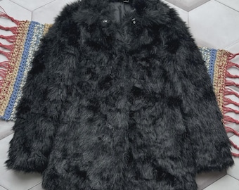 May include: A black faux fur coat with a collar and two small buttons. The coat is displayed on a patterned rug and a hexagonal tile floor. The coat has a soft, fluffy texture.