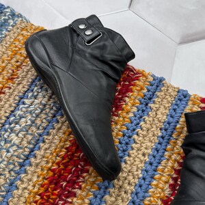 May include: Black leather ankle boot with a buckle detail. The boot is set on a colorful, textured rug with blue, red, yellow, and beige stripes. The boot has a rounded toe and a low heel.