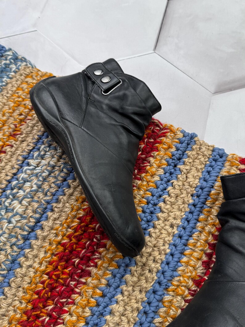 May include: Black leather ankle boot with a buckle detail. The boot is set on a colorful, textured rug with blue, red, yellow, and beige stripes. The boot has a rounded toe and a low heel.