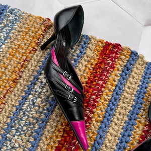 May include: A pair of black leather high heels with pink accents and multiple buckles. The heels are pointed and have a stiletto heel.