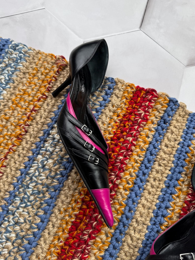 May include: A pair of black leather high heels with pink accents and multiple buckles. The heels are pointed and have a stiletto heel.