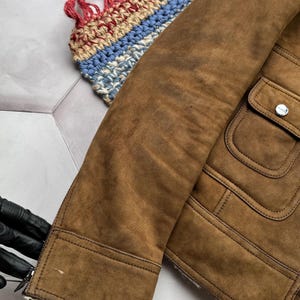 May include: Close-up of a brown suede jacket with a shearling lining. The jacket has a classic design with a buttoned pocket and a zipper closure. The sleeve is visible, showing the texture of the suede.
