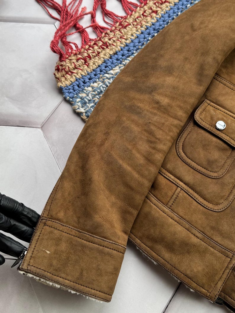 May include: Close-up of a brown suede jacket with a shearling lining. The jacket has a classic design with a buttoned pocket and a zipper closure. The sleeve is visible, showing the texture of the suede.