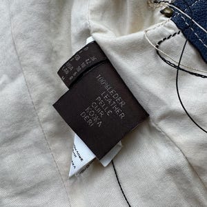以下が含まれることがあります： 茶色の革タグのクローズアップ。タグには「100% Leder Leather Pelle Cuir Кожа Deri」というテキストと、US、GB、F、CAN などのさまざまな単位で表されたサイズ情報が記載されています。