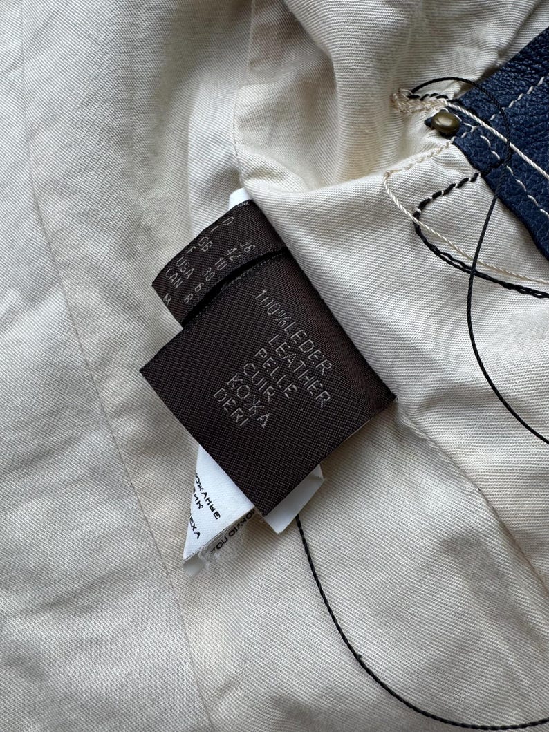 以下が含まれることがあります： 茶色の革タグのクローズアップ。タグには「100% Leder Leather Pelle Cuir Кожа Deri」というテキストと、US、GB、F、CAN などのさまざまな単位で表されたサイズ情報が記載されています。
