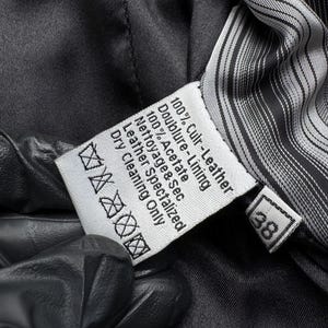 K&ouml;nnte beinhalten: Nahaufnahme eines wei&szlig;en Etiketts mit schwarzem Text, darunter "100% Culr-Leather" und "Dry Cleaning Only". Das Etikett befindet sich in einem dunklen Kleidungsst&uuml;ck mit gestreiftem Futter. Die Zahl "38" ist sichtbar.
