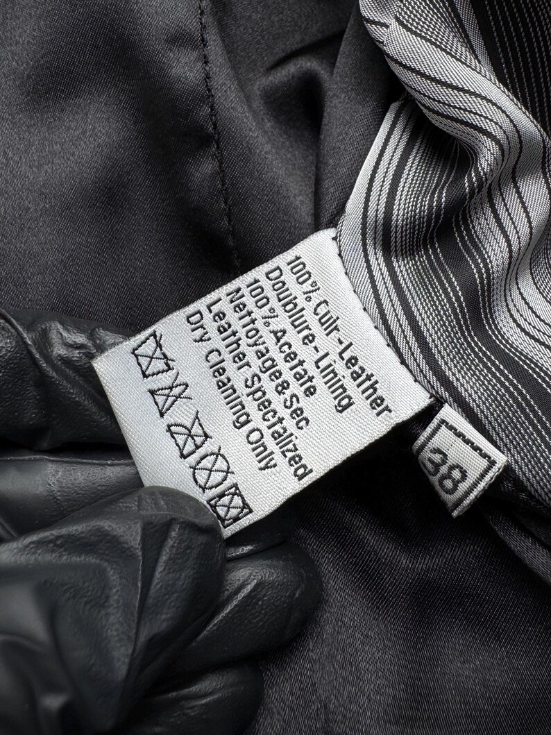 K&ouml;nnte beinhalten: Nahaufnahme eines wei&szlig;en Etiketts mit schwarzem Text, darunter "100% Culr-Leather" und "Dry Cleaning Only". Das Etikett befindet sich in einem dunklen Kleidungsst&uuml;ck mit gestreiftem Futter. Die Zahl "38" ist sichtbar.