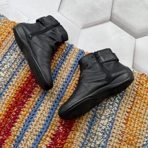 May include: A pair of black leather ankle boots with side zippers and buckle straps. The boots are placed on a colorful, striped, woven rug with blue, red, yellow, and beige hues. The background is a white hexagonal tile.