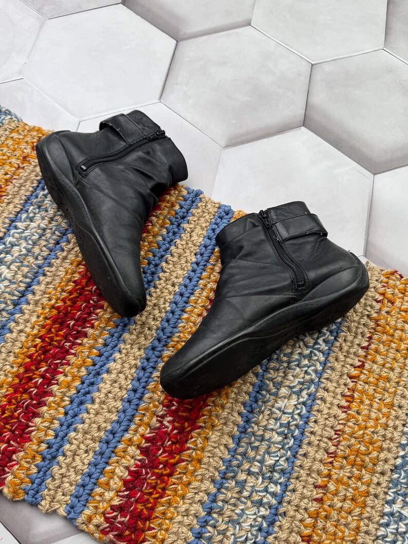 May include: A pair of black leather ankle boots with side zippers and buckle straps. The boots are placed on a colorful, striped, woven rug with blue, red, yellow, and beige hues. The background is a white hexagonal tile.