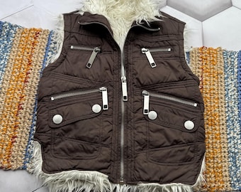 May include: A brown puffer vest with a white faux fur collar and trim. The vest has multiple zippered pockets and a zipper closure.