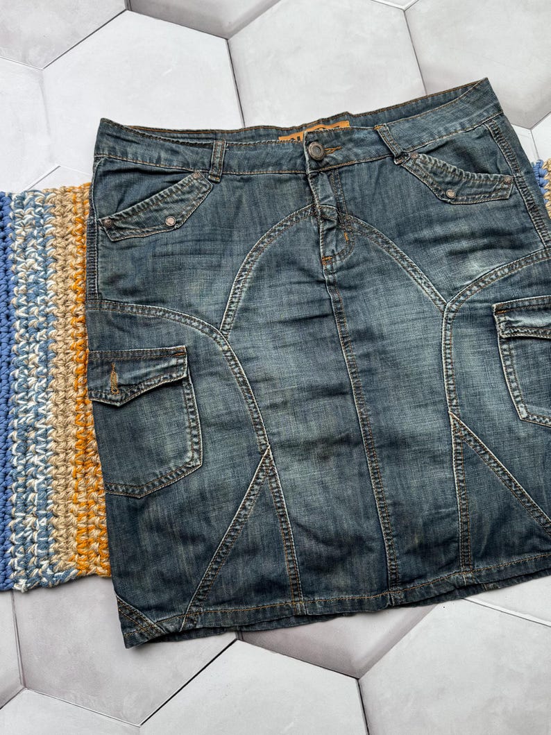 Puede incluir: Una falda vaquera con un lavado azul descolorido. La falda presenta m&uacute;ltiples bolsillos, costuras decorativas y cierre de bot&oacute;n. La falda tiene un estilo cl&aacute;sico y casual.
