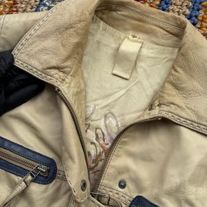 以下が含まれることがあります： 青いトリムとジッパーのディテールが付いたベージュの革のジャケット。ジャケットには、襟の内側に白いラベルが縫い付けられています。