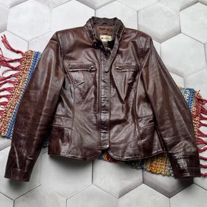 Pu&ograve; includere: Una giacca di pelle marrone con chiusura a zip e diverse tasche. La giacca ha un colletto alto e un'etichetta del marchio che recita "ADLER".