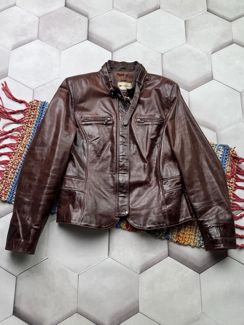 Pu&ograve; includere: Una giacca di pelle marrone con chiusura a zip e diverse tasche. La giacca ha un colletto alto e un'etichetta del marchio che recita "ADLER".
