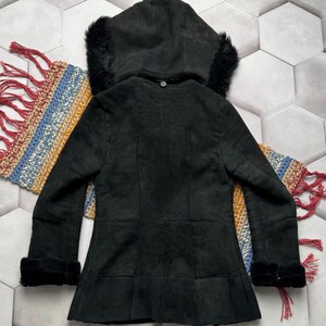 May include: Black suede hooded jacket with faux fur trim on the hood and cuffs. The jacket has a button closure and a flared hem. The back of the jacket is visible, showing the seams and the texture of the suede. The jacket is displayed on a colorful woven textile.