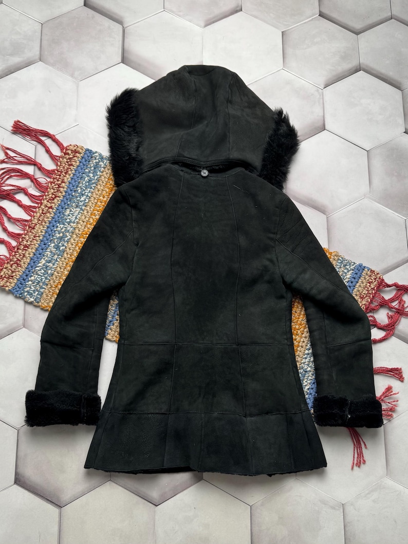 May include: Black suede hooded jacket with faux fur trim on the hood and cuffs. The jacket has a button closure and a flared hem. The back of the jacket is visible, showing the seams and the texture of the suede. The jacket is displayed on a colorful woven textile.