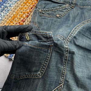 Puede incluir: Primer plano de unos pantalones cortos vaqueros azules con m&uacute;ltiples bolsillos y costuras decorativas. Los pantalones cortos tienen un lavado descolorido y cierre de bot&oacute;n. Los pantalones cortos se muestran sobre una superficie colorida y estampada.