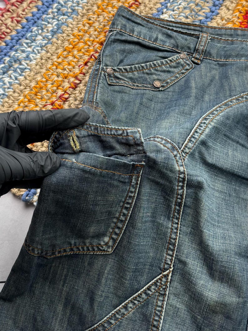 Puede incluir: Primer plano de unos pantalones cortos vaqueros azules con m&uacute;ltiples bolsillos y costuras decorativas. Los pantalones cortos tienen un lavado descolorido y cierre de bot&oacute;n. Los pantalones cortos se muestran sobre una superficie colorida y estampada.