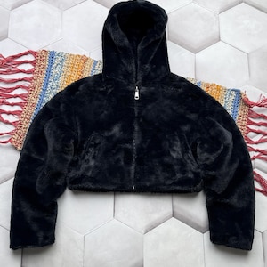 May include: A black faux fur hooded jacket with a silver zipper closure. The jacket has long sleeves and a cropped length. The soft, plush material creates a cozy look. The jacket is displayed on a patterned surface.