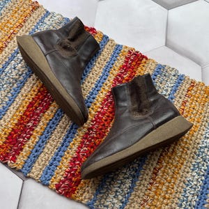 May include: A pair of brown leather ankle boots with a low wedge heel. The boots have a textured collar and are set on a colorful striped rug. The boots are a neutral color and could be worn with many outfits.