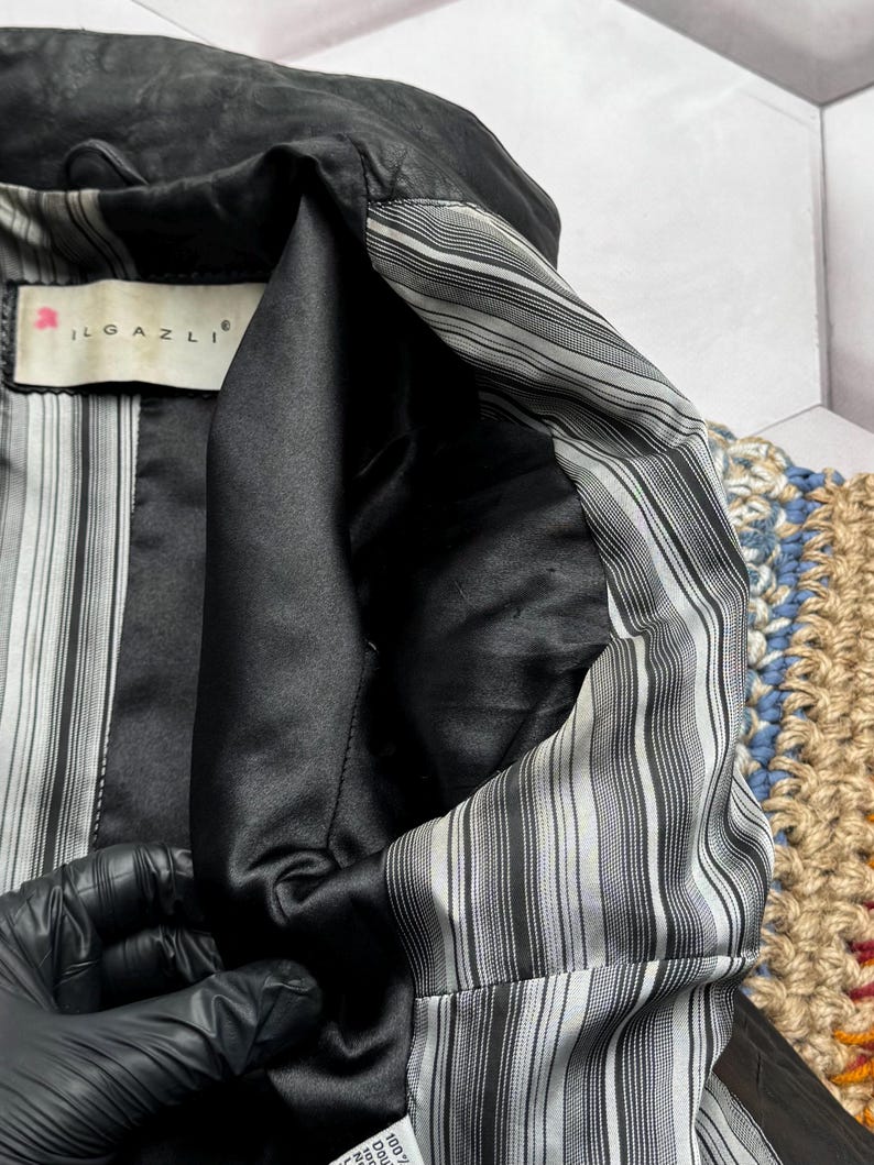 K&ouml;nnte beinhalten: Nahaufnahme einer schwarzen Lederjacke mit gestreiftem Innenfutter. Das Jackeninnere besteht aus einem schwarzen, satinartigen Material und tr&auml;gt das Etikett "ILGAZLI". Das Futter hat d&uuml;nne, vertikale grau-wei&szlig;e Streifen.