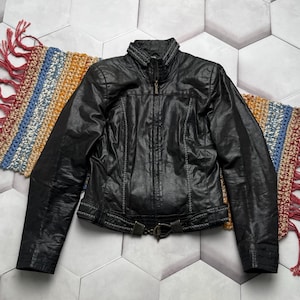 K&ouml;nnte beinhalten: Schwarze Lederjacke mit Rei&szlig;verschluss, dekorativen N&auml;hten und einem G&uuml;rtel in der Taille. Die Jacke hat einen Stehkragen und wird auf einem gemusterten Teppich pr&auml;sentiert. Ein klassisches Kleidungsst&uuml;ck.