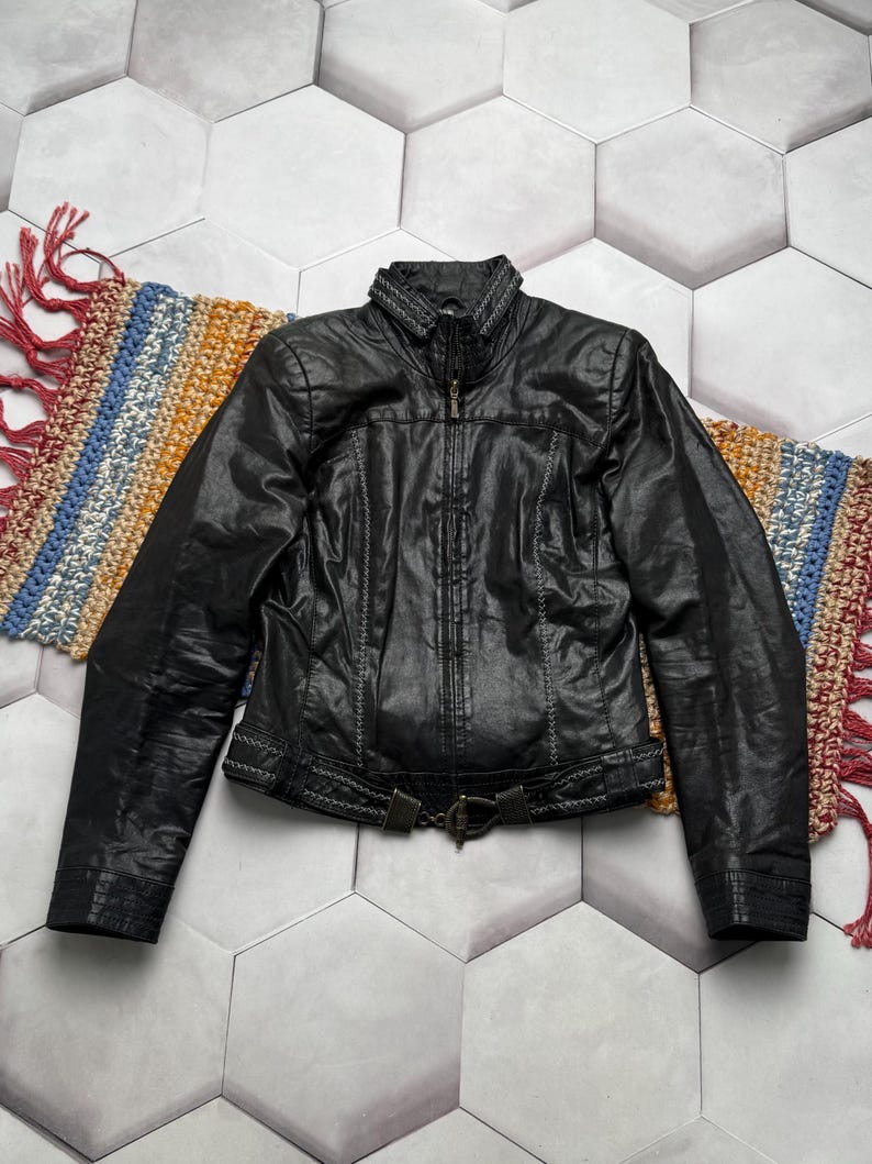 K&ouml;nnte beinhalten: Schwarze Lederjacke mit Rei&szlig;verschluss, dekorativen N&auml;hten und einem G&uuml;rtel in der Taille. Die Jacke hat einen Stehkragen und wird auf einem gemusterten Teppich pr&auml;sentiert. Ein klassisches Kleidungsst&uuml;ck.