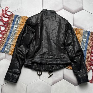 K&ouml;nnte beinhalten: Schwarze Lederjacke mit strukturierter Oberfl&auml;che, Stehkragen und dekorativen N&auml;hten am Bund. Die Jacke hat einen Rei&szlig;verschluss und einen klassischen Biker-Stil. Die Jacke wird auf einem farbenfrohen Webteppich pr&auml;sentiert.