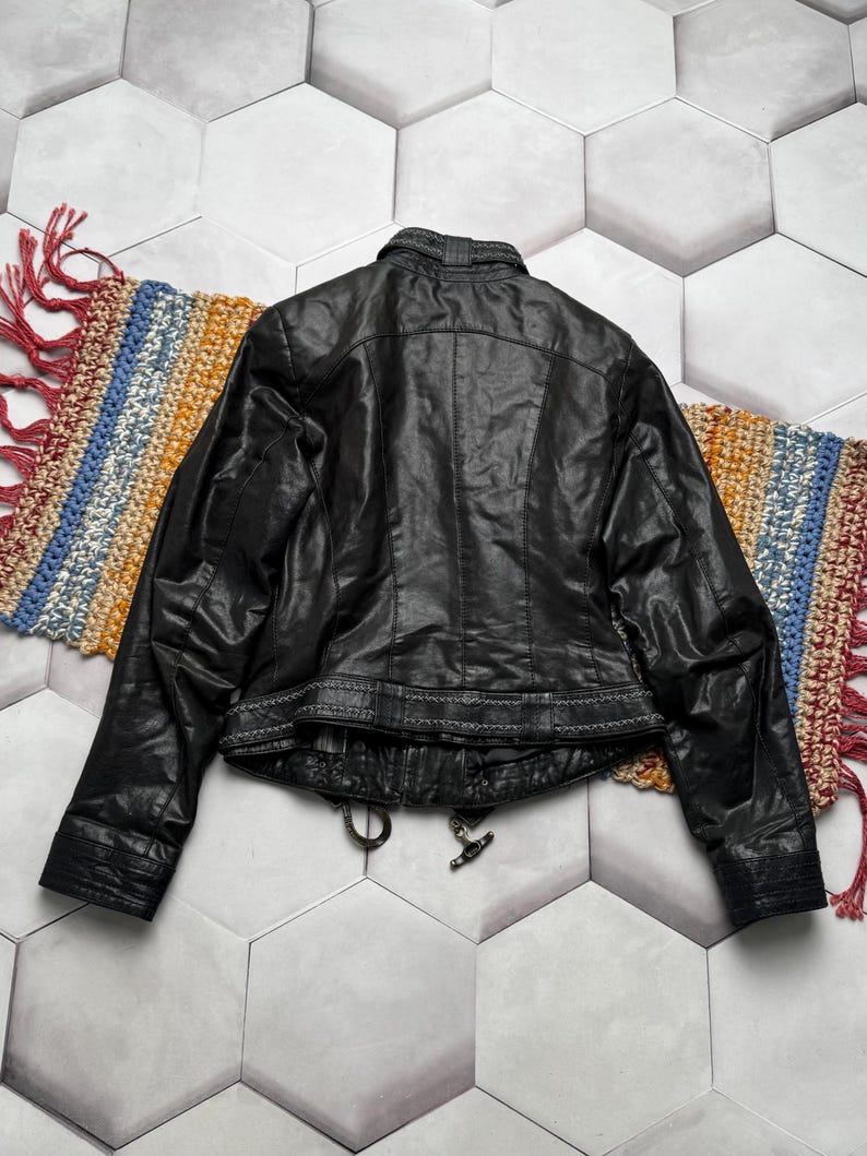 K&ouml;nnte beinhalten: Schwarze Lederjacke mit strukturierter Oberfl&auml;che, Stehkragen und dekorativen N&auml;hten am Bund. Die Jacke hat einen Rei&szlig;verschluss und einen klassischen Biker-Stil. Die Jacke wird auf einem farbenfrohen Webteppich pr&auml;sentiert.