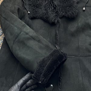 May include: Black shearling coat with a plush, furry collar and cuffs. The coat features a button closure and a soft, suede-like texture. The sleeves are long and the coat appears warm and cozy.