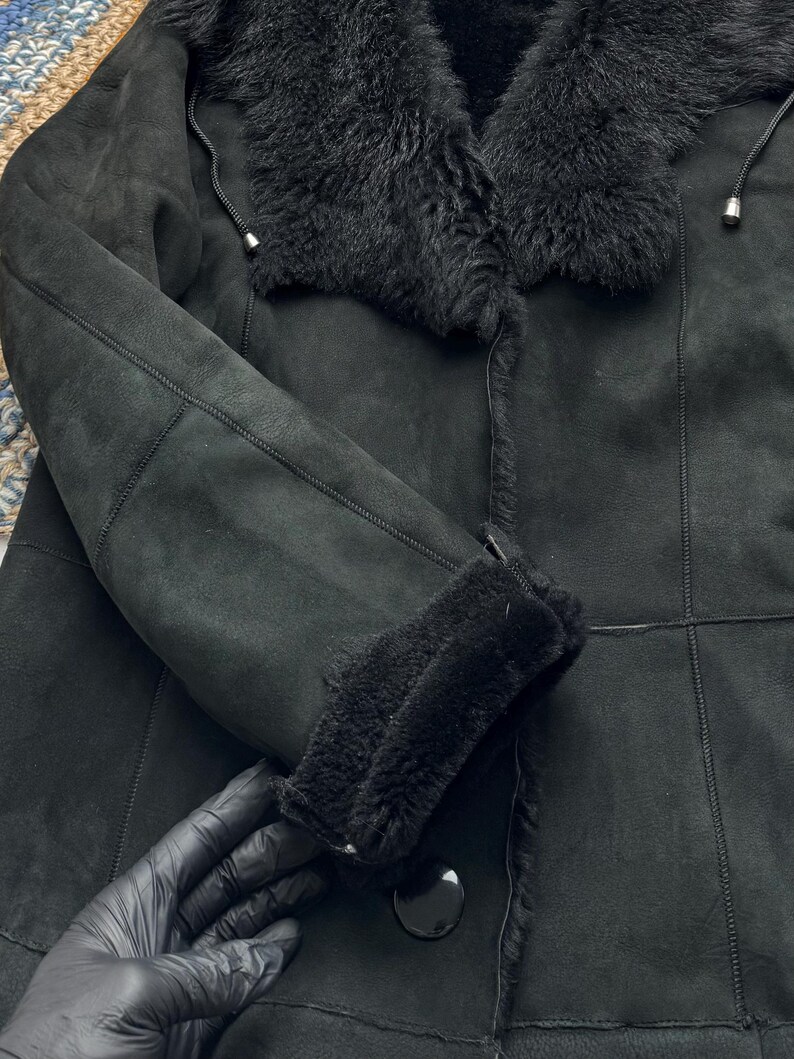 May include: Black shearling coat with a plush, furry collar and cuffs. The coat features a button closure and a soft, suede-like texture. The sleeves are long and the coat appears warm and cozy.