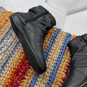 May include: Black leather ankle boots with side zippers and a strap detail. The boots are set on a colorful, striped rug with blue, red, and tan hues. The boots have a low heel.