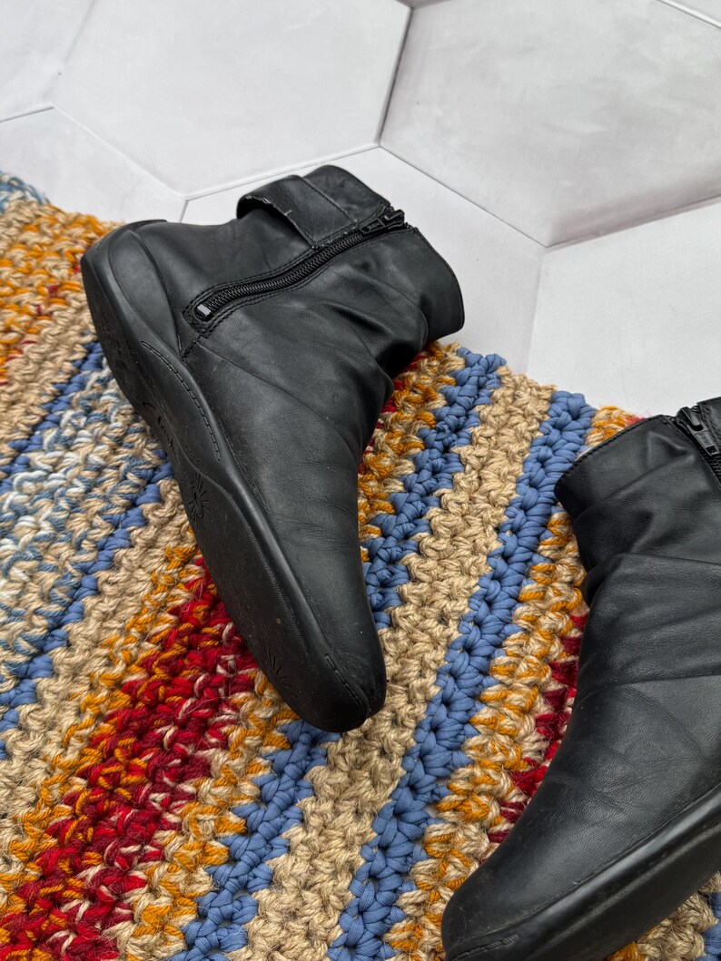 May include: Black leather ankle boots with side zippers and a strap detail. The boots are set on a colorful, striped rug with blue, red, and tan hues. The boots have a low heel.
