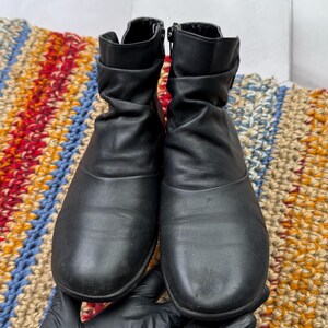 May include: A pair of black ankle boots with a scrunched design. The boots have zippers on the sides and are made of a smooth material. The boots are displayed on a colorful, patterned rug.