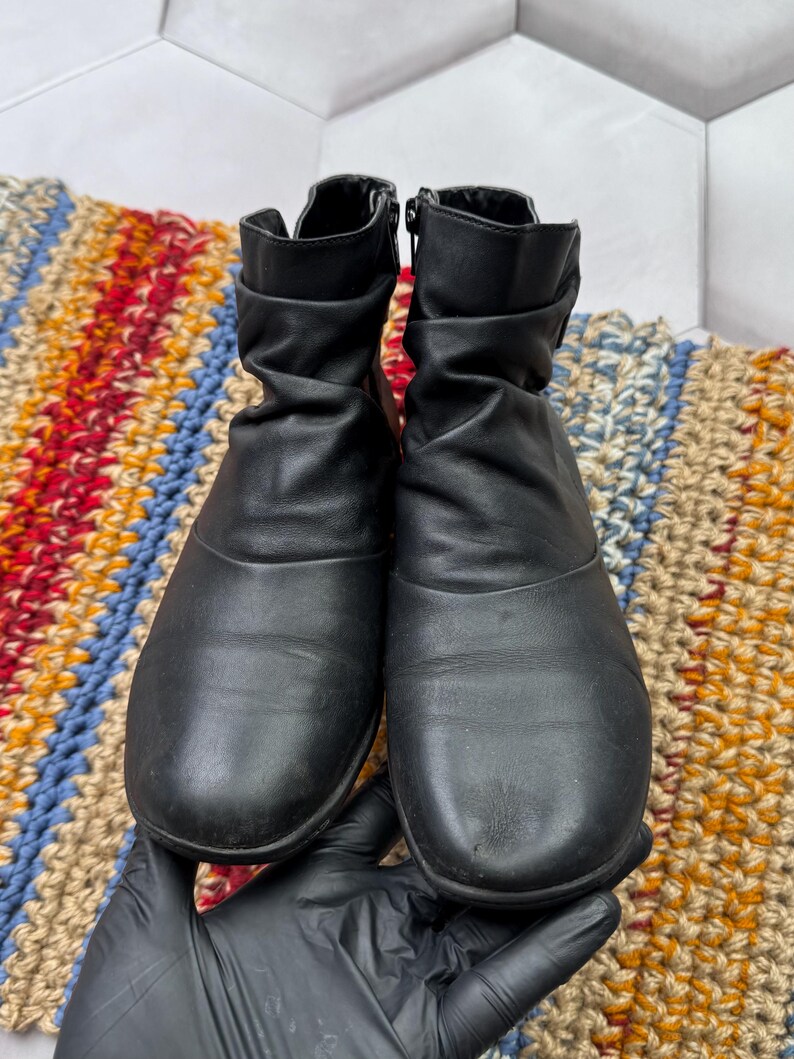 May include: A pair of black ankle boots with a scrunched design. The boots have zippers on the sides and are made of a smooth material. The boots are displayed on a colorful, patterned rug.