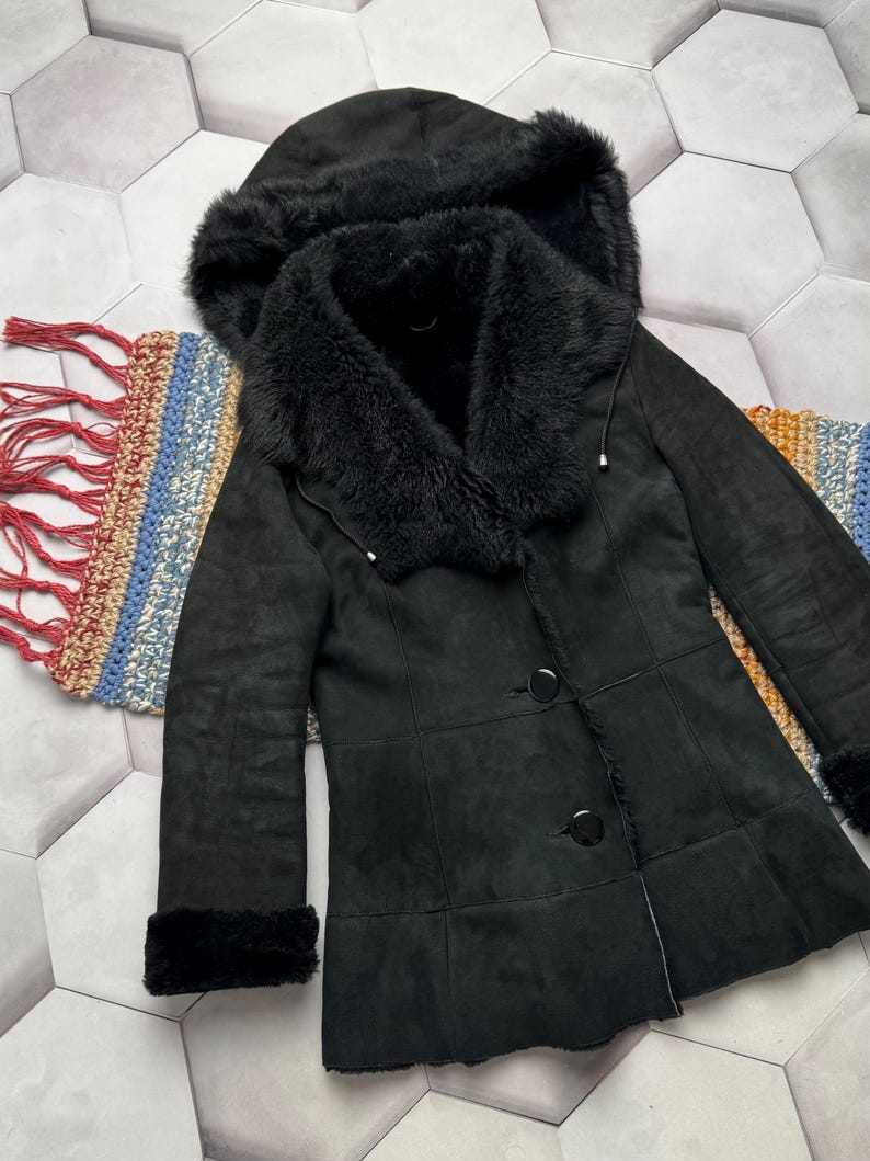 May include: A black coat with a hood and faux fur trim. The coat has a button closure and fur trim on the cuffs. The coat is made of a suede-like material. The coat is displayed on a patterned surface.
