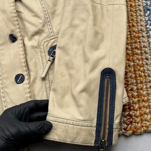 以下が含まれることがあります： 青いジッパーのディテールとステッチが付いたベージュの革のジャケット。ジャケットはボタン留めと襟があります。